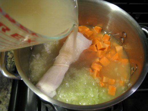 pouring broth into large stainless steel soup pot with onion, celery, carrot and herb bundle