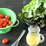 garlic salad dressing in a small glass canister on a wooden board with cherry tomatoes in a green colander, a head of red leaf lettuce and a small whisk