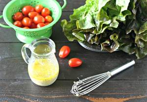 garlic salad dressing in a small glass canister on a wooden board with cherry tomatoes in a green colander, a head of red leaf lettuce and a small whisk