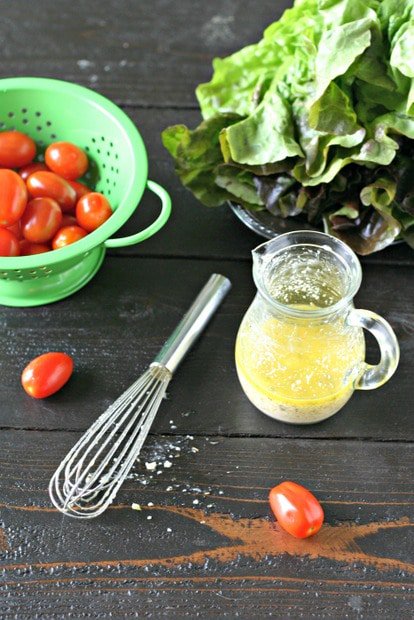 garlic salad dressing in a small glass canister on a wooden board with cherry tomatoes in a green colander, a head of red leaf lettuce and a small whisk on a dark wood background