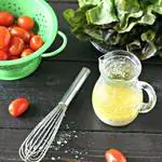 garlic salad dressing in a small glass canister on a wooden board with cherry tomatoes in a green colander, a head of red leaf lettuce and a small whisk