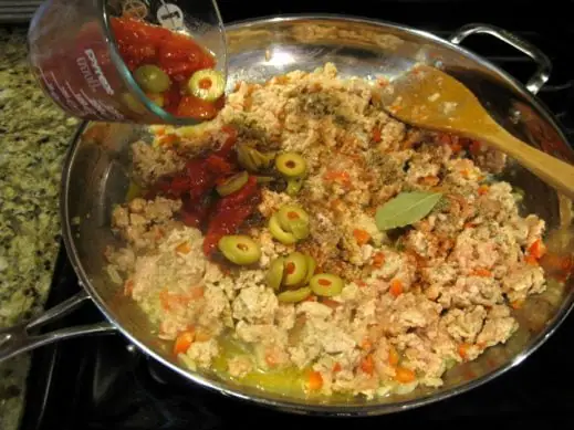 adding spices, olives and tomatoes to chicken and veggies for picadillo