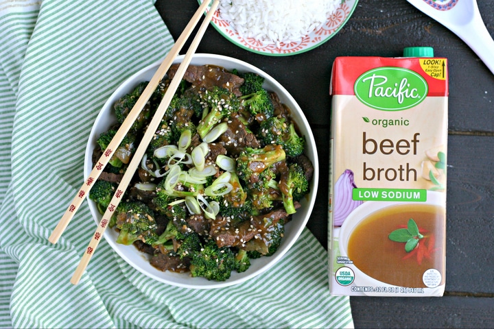 a bowl of beef and broccoli on a green and white striped linen with chopsticks, rice and a carton of beef broth