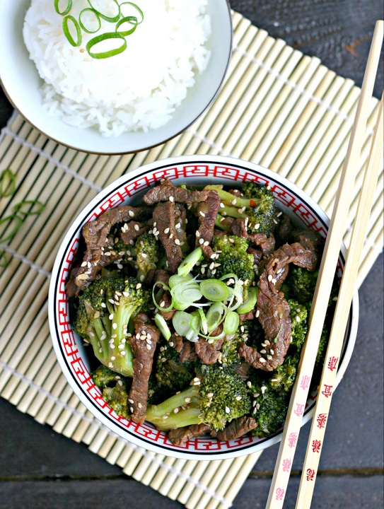 bamboo mat with bowl of beef and broccoli, chopsticks resting on bowl, small bowl of white rice with green scallions on top