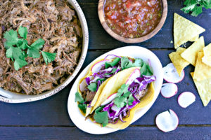 bowl of pulled pork made in the pressure cooker, tacos and salsa with sliced radish and chips