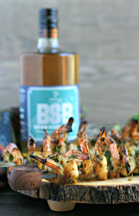 close up of grilled shrimp skewers on a wood platter with bottle of bourbon in background