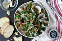 a white bowl of steamed clams in a spicy tomato sauce with crusty bread, red pepper flakes and a bowl for the shells