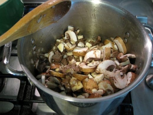 stainless steel soup pot on stovetop, using a wooden spoon to stir mushrooms, parsnips and onion into meat mixture