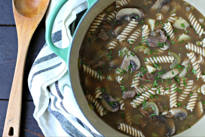 dutch oven filled with beef noodle soup sitting on a kitchen towel with a large wooden spoon