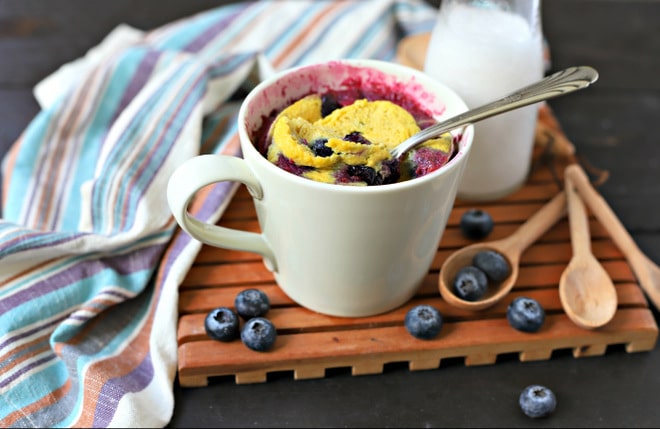 blueberry mug cake with silver spoon sticking out surrounded by fresh blueberries, a glass of milk and a napkin on a slotted wood tray