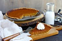 slice of pumpin pie topped with whipped cream on a wood cutting board in front of whole pie with glass of milk in glass jar