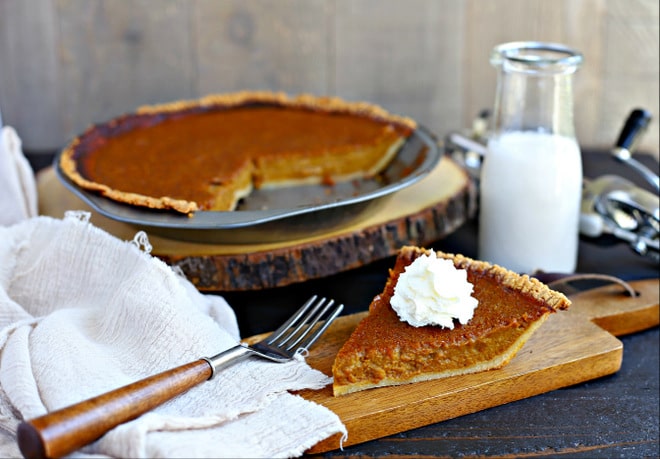 slice of pumpin pie topped with whipped cream on a wood cutting board in front of whole pie with glass of milk in glass jar
