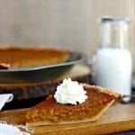 close up of slice of pumpin pie topped with whipped cream on a wood cutting board in front of whole pie with glass of milk in glass jar