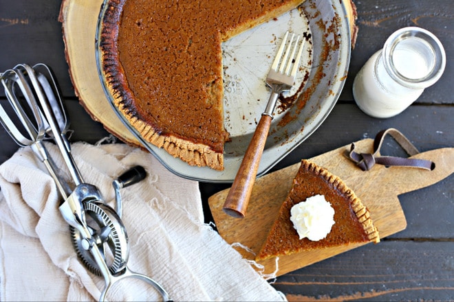 overhead view of slice of pumpin pie topped with whipped cream on a wood cutting board in front of whole pie with glass of milk in glass jar