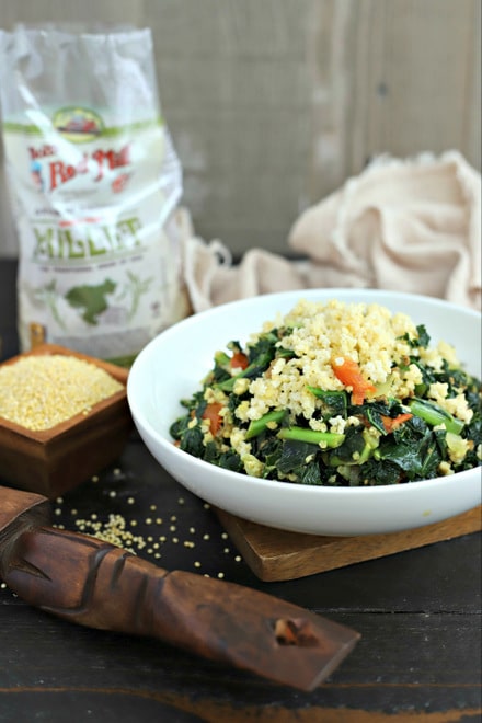 white bowl with cooked sukuma wiki topped with millet next to small square wood bowl of uncooked millet and african wood serving spoon plus an open bag of millet and distressed linen