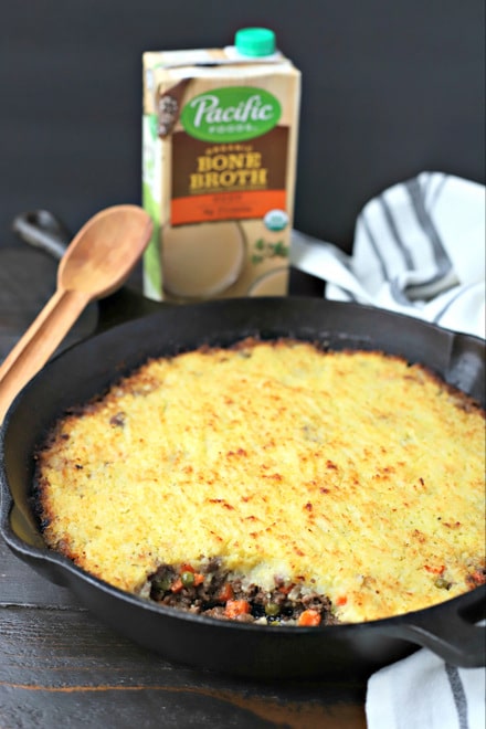 birds eye view of cast iron skillet filled with cooked easy shepherds pie and wooden spoon. in front of a carton of Pacific Foods Beef Bone Broth and a black and white linen