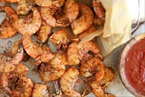 close up overhead shot of cooked old bay shrimp topped with extra old bay seasoning with wood bowl filled with cocktail sauce