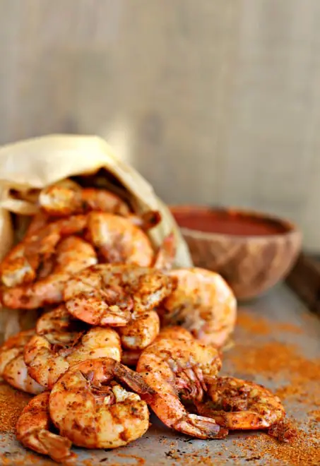 cooked old bay shrimp falling out of a paper bag in front of cocktail sauce on a tray sprinkled with old bay