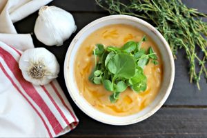 rustic bowl filled with creamy garlic soup topped with watercress leaves. on wooden table with fresh thyme, whole heads of garlic and a red and white linen