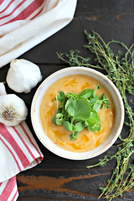 rustic bowl filled with creamy garlic soup topped with watercress leaves. on wooden table with fresh thyme, whole heads of garlic and a red and white linen