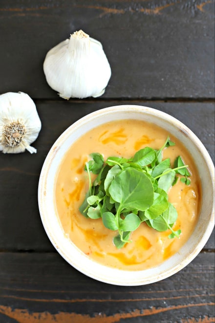 white bowl of creamy garlic soup topped with watercress leaves next to two whole heads of garlic