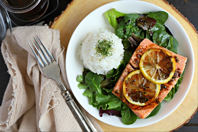 white plate with bed of mixed greens, cooked salmon topped with sliced lemon and ball of white rice topped with spinach seasoning. balsamic dressing drizzled on top of salmon and salad