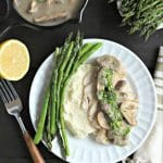 white dinner plate with asparagus, mashed cauliflower and poached chicken breast with creamy mushroom sauce alongside a cut lemon, flatware with wood handles and a small cast iron pot of more mushroom sauce