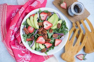 white bowl with strawberry salad and jar of balsamic dressing next to wood serving spoons and red linen