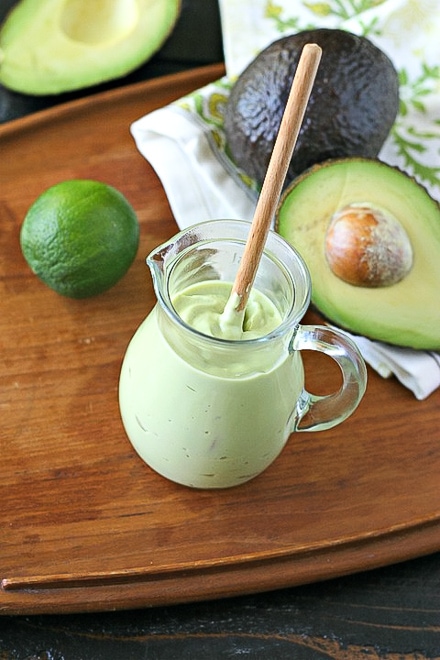 vegan avocado crema in glass jar on a wood tray with cut avocado and lime
