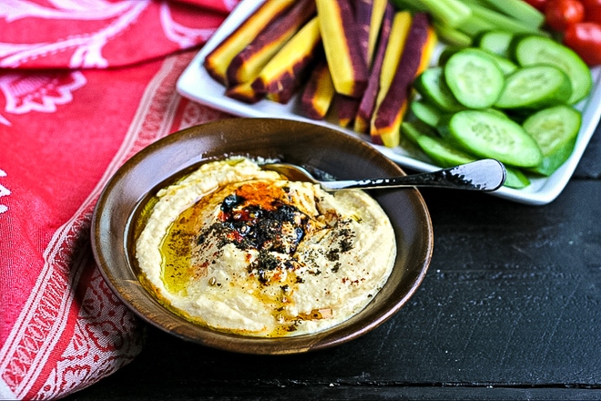 wood bowl of hummus topped with spices and olive oil in front of tray of vegetables