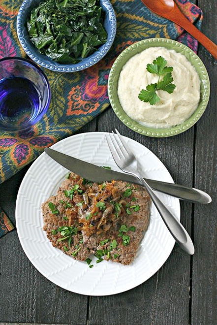 overhead of dinner table with cube steak on a white plate, mashed cauliflower, and greens