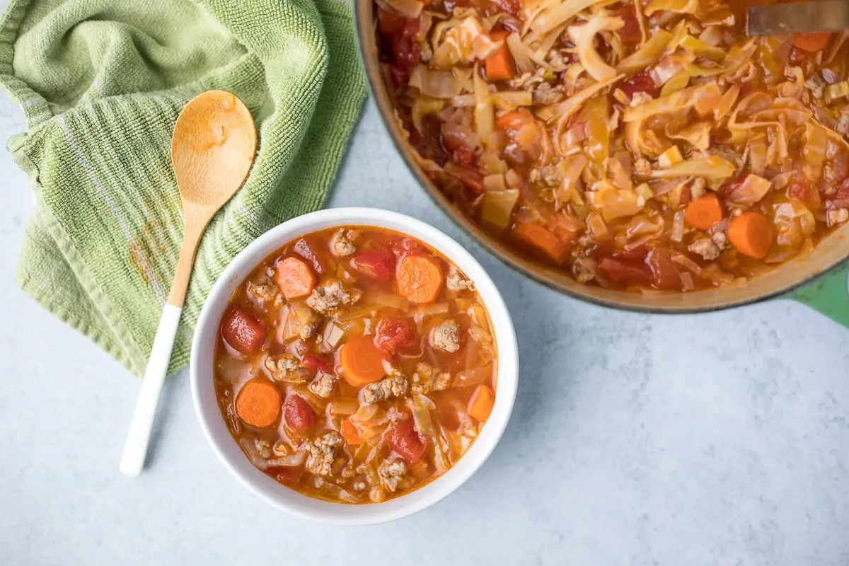 white bowl of sausage and cabbage soup next to a large pot of the same soup