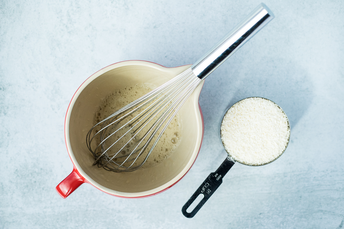 mixing bowl with egg whites and whisk next to measuring cup of coconut
