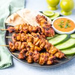 gray plate with cooked chicken kabobs, sliced cucumber, spicy peanut sauce, cut lime, and pita bread
